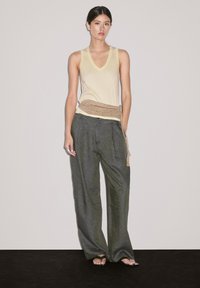 Ein Model steht vor einem schlichten Hintergrund und trägt ein leichtes Tanktop, weite Hosen und einen dekorativen beigen Gürtel, der einen stilvollen Look unterstreicht.
