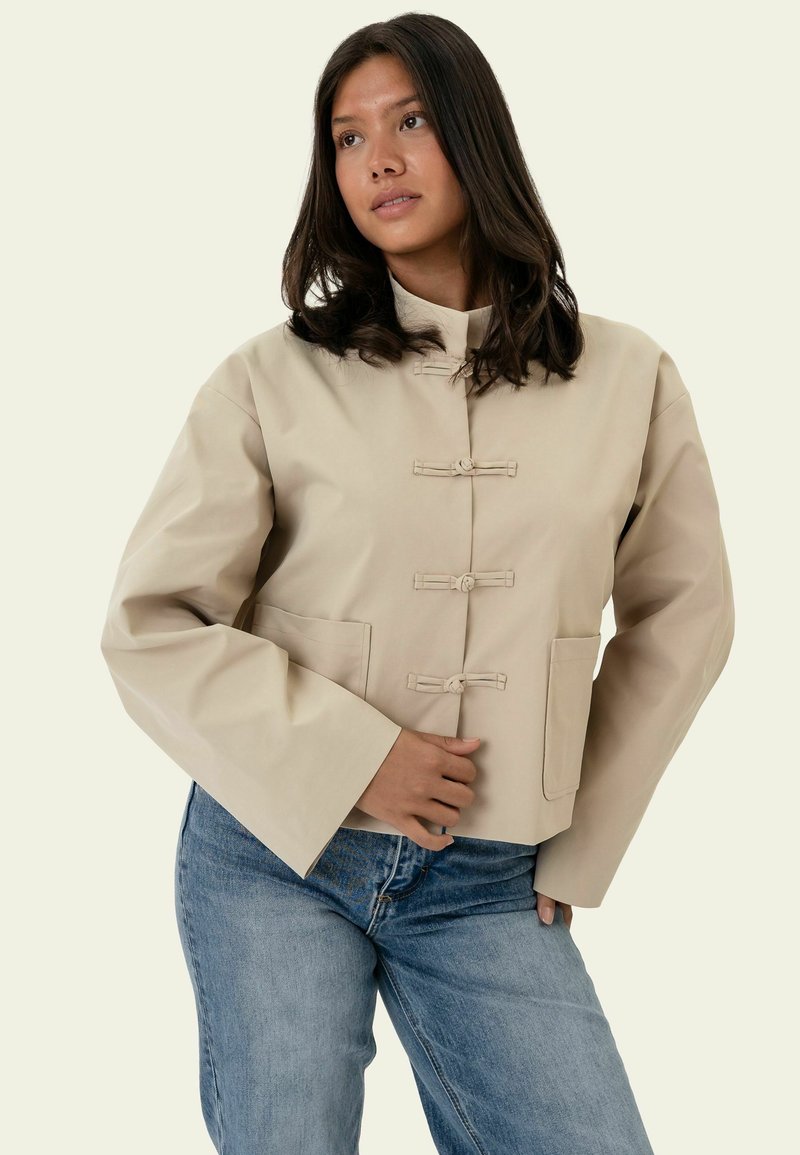 Femme aux cheveux mi-longs foncés portant une veste beige boutonnée et un jean bleu, debout avec une main posée sur la hanche.