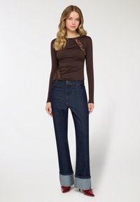 Top marrone a maniche lunghe con dettagli in pizzo, abbinato a jeans blu scuro a gamba larga con risvolti. Accessori inclusi: calzature rosse.