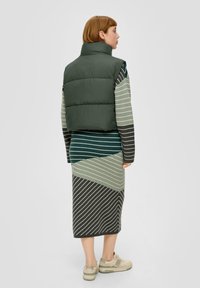 Grüne, puffige Weste über einem mehrfarbigen, gestreiften Kleid mit diagonalen Mustern in Grün, Grau und Schwarz. Beigefarbene Sneaker runden das Outfit ab.