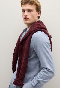 Maglione bordeaux a trecce appoggiato su una camicia azzurra chiusa con bottoni, caratterizzato da un motivo testurizzato e polsini a coste.