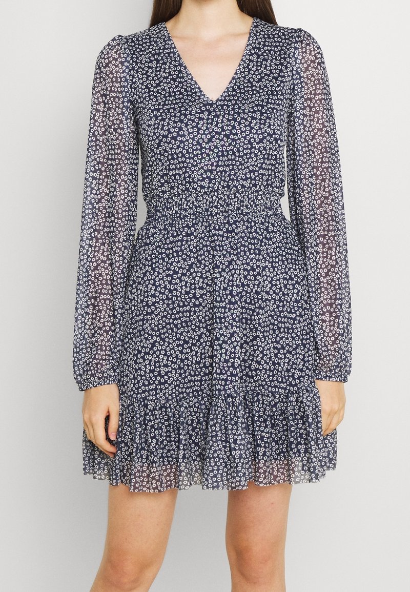 Femme portant une robe fleurie bleu marine avec des manches longues transparentes, un col en V, une taille élastique et un ourlet à volants, debout sur un fond neutre.