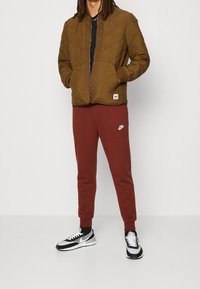 Brun vadderad jacka med ribbade manschetter, tillsammans med vinröda sweatpants med en vit logotyp, och svart-vita sneakers.
