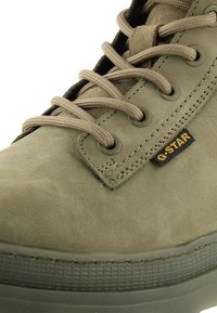 Gros plan d'une basket vert olive à lacets avec une semelle texturée et une étiquette de marque "G-STAR" visible sur le côté.