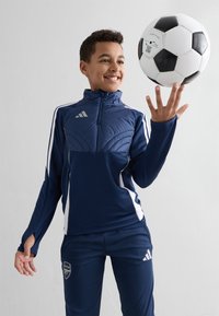 Chlapec v námořní modrej športovej súprave točí čierno-bielym futbalovým loptou na svojom prstovom konci, usmieva sa proti jednoduchému svetlému pozadiu.