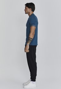 Camiseta azul ajustada de manga corta, con un logo en el brazo, combinada con pantalones de chándal negros y zapatillas blancas, de pie de lado.