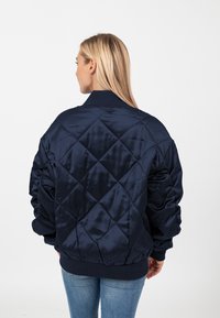 Marineblauwe gewatteerde pufferjack met een satijnen afwerking. Kenmerkt ribgebreide manchetten en kraag, en heeft een ontspannen pasvorm. Zichtbaar ruitpatroon op de achterkant.