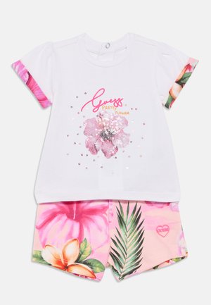 Tøyskrud til småbarn med hvit kortermet topp som har en rosa paljettblomst og teksten «Guess Pretty Flower», kombinert med rosa blomstrete shorts.