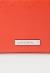 Jasnopomarańczowy portfel skórzany o fakturowanej powierzchni z metalową płytką w kolorze srebrnym, na której widnieje napis "KARL LAGERFELD." Charakteryzuje się eleganckim, minimalistycznym designem.