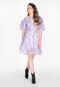 Blomstret kjole i lilla med puffede ærmer og udskæringer, med et lagdelt kant. Båret med sorte ankelstøvler. Glat, let stof.