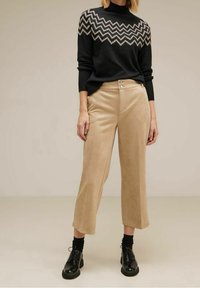 Kvinna klädd i en svart och grå zigzag-mönstrad tröja, beige capribyxor med knapprar, svarta strumpor och svarta snörskor.