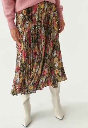 Frau trägt einen plissierten, geblümten Midirock mit weißen Absatzstiefeln und einem pinken Pullover, gezeigt von der Taille bis zu den Füßen auf weißem Hintergrund.