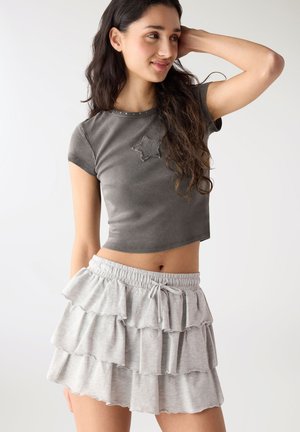 Giovane donna con i capelli lunghi e scuri che indossa una t-shirt grigia corta con dettaglio a stella e una minigonna a balze a strati grigio chiaro.