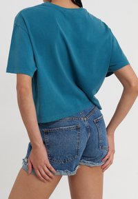 Camiseta de algodón en color teal con mangas cortas, corte relajado y cuello redondo, combinada con pantalones cortos de denim desgastado con dobladillos deshilachados y bolsillos.