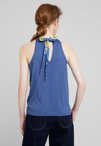 Modrý bezrukávový top s klíčovou dírkou na zádech, zdobený květinovým vzorem na krčním šátku. Měkká tkanina a uvolněný střih.