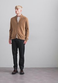 Tânăr stând în interior, purtând un pulover cu fermoar maro, un tricou alb, pantaloni negri și pantofi de dressing maro, în fața unei pereți gri uni.