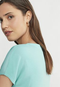 Kortärmad mintgrön topp med rund halsringning och mjuk textur, tillsammans med ett guldörhänge som har en jämn finish.