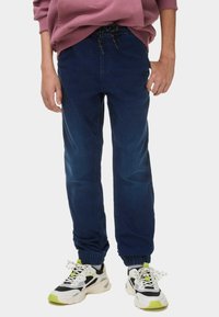 Pantalones jogger azul marino con cinturilla elástica y cordón. La tela tiene una textura suave. Se combinan con zapatillas blancas y negras con estampado.