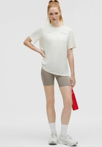Junge Frau in weißem Sportshirt und beigen Shorts, die eine rote Wasserflasche hält, vor einem schlichten grauen Hintergrund stehend.