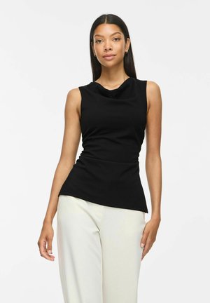 Femme aux longs cheveux foncés porte un haut noir sans manches et un pantalon clair, debout devant un fond clair uni.