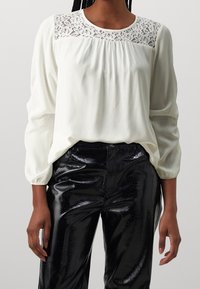 Blusa bianca con dettagli in pizzo sul colletto e parte anteriore plissettata, abbinata a pantaloni neri in ecopelle lucida con finitura testurizzata.