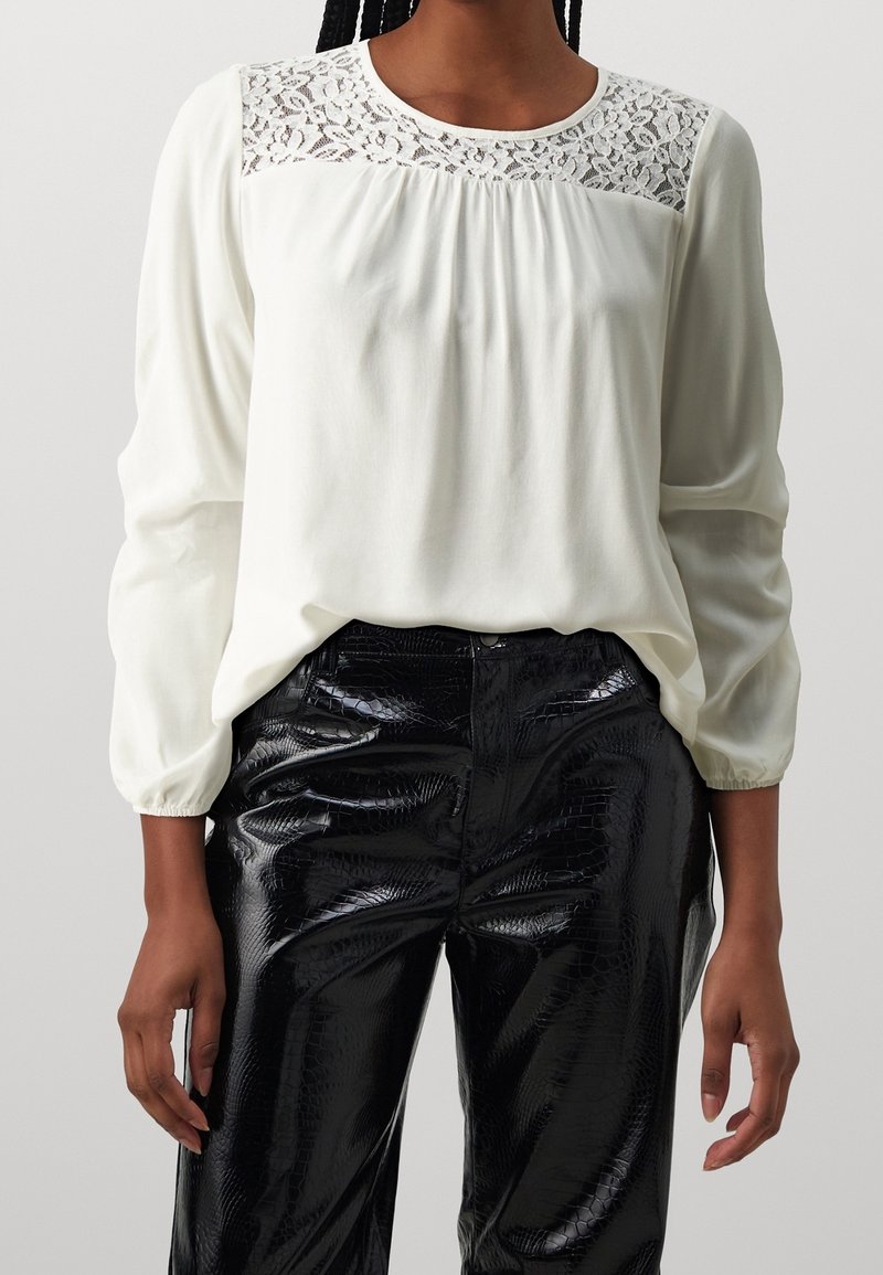 Blusa bianca con dettagli in pizzo sul colletto e parte anteriore plissettata, abbinata a pantaloni neri in ecopelle lucida con finitura testurizzata.