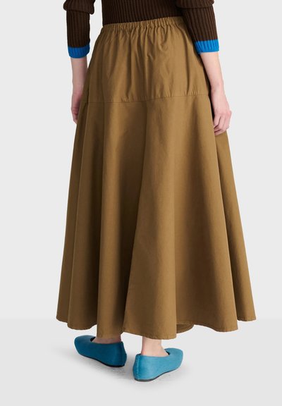 Personne portant une jupe longue marron, un pull marron avec des poignets bleus et des chaussures plates bleu vif, debout sur un fond clair.