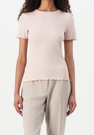 Nainen pukeutuneena vaaleanpunaiseen lyhythihaiseen resoripaitaan, jossa on salaattireunaiset helmassa, yhdistettynä beigeihin räätälöityihin housuihin.