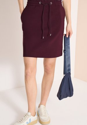 Femme portant une jupe bordeaux à cordon, des baskets blanches, tenant un sac à main bleu marine par sa bandoulière à son côté.