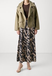 Chaqueta de piel sintética verde oliva con cremalleras, combinada con una falda maxi de patrón negro y beige y sandalias de tacón transparente.