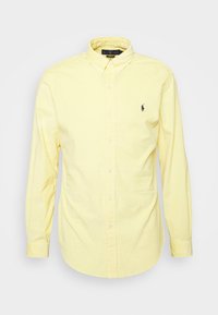 Žlutá košile s dlouhým rukávem z bavlny, s límečkem na knoflíky a malým námořnickým logem na levé straně hrudi. Hladká textura, úzký střih.