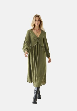 Mujer rubia caminando con un vestido largo verde oliva de mangas abullonadas y botas negras hasta la rodilla, llevando un collar de cadena dorada.