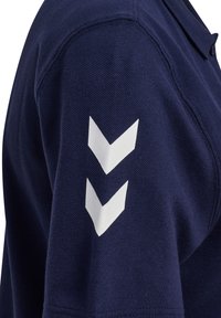 Marineblå poloshirt med tekstureret stof, der har et hvidt-chevronmønster på venstre ærme. Klassisk krave og knaplukning.