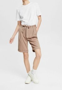 Pantalones cortos beige a medida con un cinturón atado, dobladillo enrollado, combinados con una camiseta blanca lisa y zapatillas blancas. Textura de tela suave.