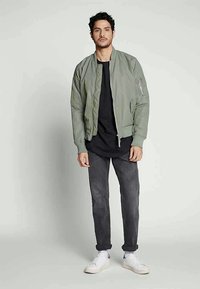 Ανοιχτό πράσινο bomber jacket με ριγωτές μανσέτες και στρίφωμα, μπροστινό φερμόυρ, και πλάγιες τσέπες, συνδυασμένο με μαύρη μπλούζα και γκρι τζιν. Λευκά αθλητικά παπούτσια.