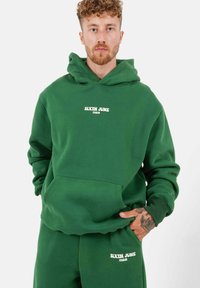 Felpa verde con tasca a marsupio, caratterizzata dal testo bianco "SIXTH JUNE PARIS". Tessuto morbido, vestibilità comoda, cappuccio spesso, polsini a coste. Pantaloni coordinati.