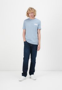 Hellblaues Baumwoll-T-Shirt mit einer Brusttasche, kombiniert mit dunkelblauen Jeans und weißen Sneakern, vor einem schlichten Hintergrund stehend.