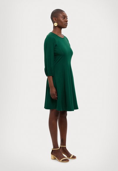 Robe verte longueur genoux avec des manches bouffantes trois quarts, en tissu lisse, accompagnée de sandales à talons blocs couleur beige.