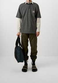 Pasiasta czarno-szara koszulka polo z naszywką z logo, na białej koszuli z długim rękawem. Oliwkowe spodnie cargo i ciemne sandały, trzyma czarną torbę.