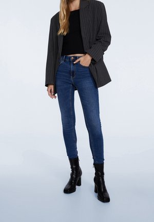 Blazer grigio scuro a righe sottili sopra un maglione cropped nero, abbinato a jeans skinny blu e stivali alla caviglia neri con tacco. Tessuti morbidi.