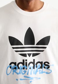 Bílý bavlněný tričko s černým logem Adidas a modrým textem "ORIGINALS". Má kulatý výstřih a krátké rukávy. Hladká textura.