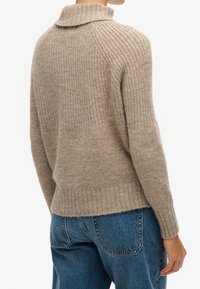 Person wearing a ribbed beige sweater with a collar and blue jeans, shown from the back against a plain white background.