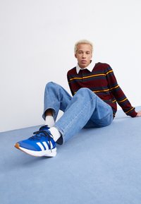 Tênis atléticos azuis com listras brancas, usados com jeans de um tom de azul claro e meias brancas. A camisa é bordô com listras horizontais em amarelo e azul.