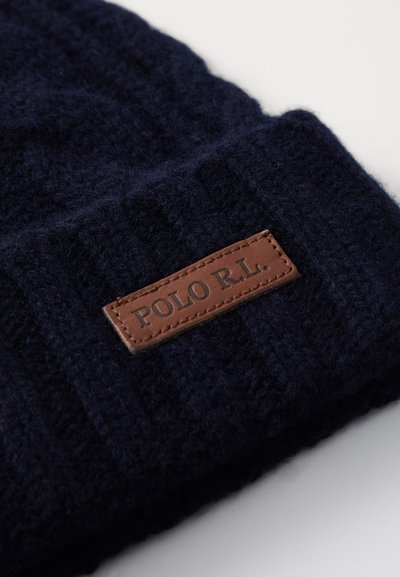 Gorro de malha azul-marinho com textura canelada e um patch de couro castanho rotulado "POLO R.L." na parte da frente. Tecido suave e quente.