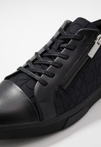 Černé tenisky s texturovaným svrškem s opakujícím se vzorem loga, hladkými koženými akcenty, kulatou špičkou a plochými tkaničkami.