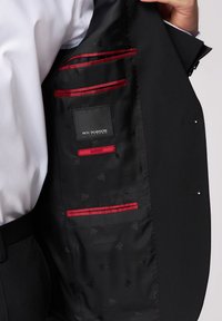 Interno di giacca nera con fodera nera a motivo. Presenta dettagli rossi, diverse tasche e un'etichetta con il marchio.