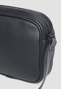 Schwarze Leder-Umhängetasche mit strukturierter Oberfläche, ausgestattet mit einem Reißverschluss oben, flacher Form und verstellbarem Tragegurt.