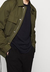 Kurtka typu bomber w oliwkowozielonym kolorze z przednimi kieszeniami, zamkiem błyskawicznym i ściągaczami przy mankietach, noszona na ciemnogranatowym swetrze z okrągłym dekoltem i czarnych spodniach.