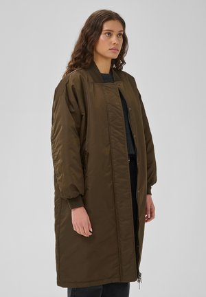 Femme aux longs cheveux bouclés portant un long manteau matelassé vert olive par-dessus des vêtements sombres, debout devant un fond clair uni.