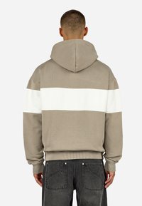 Φούτερ με κουκούλα σε ταμπά απόχρωση με λευκή οριζόντια ρίγα στο πίσω μέρος. Φτιαγμένο από υφασμάτινο βαμβάκι, διαθέτει ριμπ μανσέτες και τελείωμα.